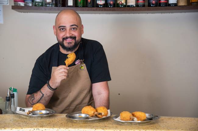 Homem segurando coxinha mordida atrás de balcão de bar vestindo avental bege com vasilhas de inox com outras coxinhas no balcão.