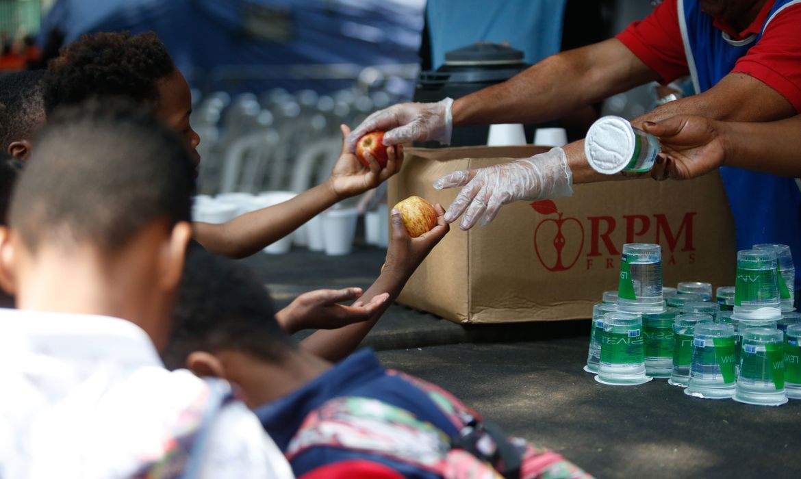 a imagem mostra as mãos de uma criança negra recebendo maçãs. a mes acom itens a serem distribuídos mostra também copos plásticos de água.
