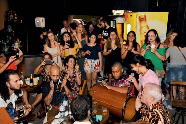 Roda de samba em primeiro plano e, ao fundo, público em pé batendo palmas dentro de bar