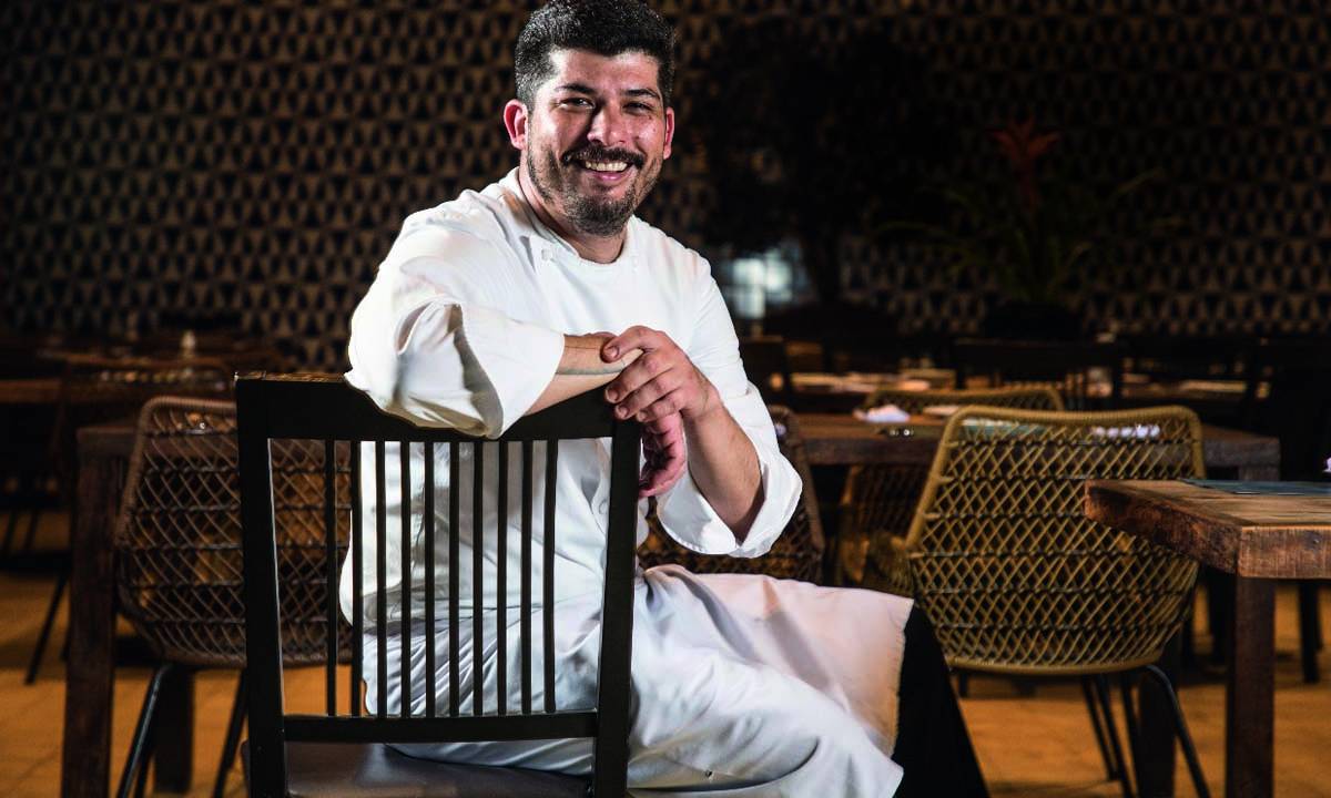 A imagem mostra o chef José Guerra Netto dentro de um salão de restaurante com mesas e cadeiras ao fundo. Ele está sentado em uma cadeira de madeira, usando o avental branco e sorrindo para a foto.
