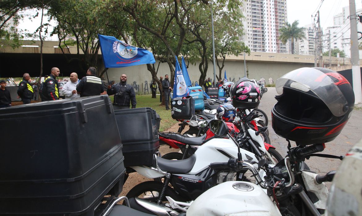 motos paradas e motoboys de pé. um deles segura uma bandeira com o logo do sindicato.