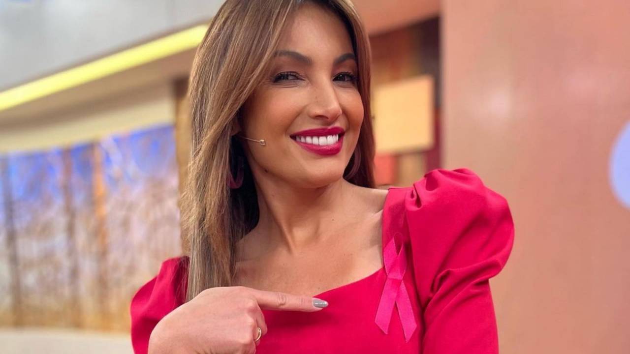 Mulher branca de cabelos longos posa sorrindo com um dedo indicador exibindo a roupa rosa.