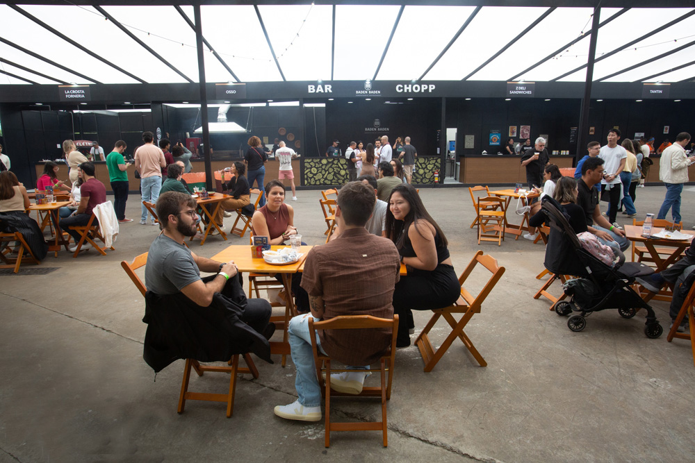 Área externa com várias mesas e cadeiras de madeira, pessoas estão sentadas conversando. Ao fundo é possível ver um estande preto.