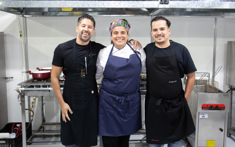 FEIRA DOS CAMPEÕES COMER E BEBER 2023 Três chefs lado a lado sorriem para a foto. O da esquerda é Kazuo Harada, que tem cabelos escuros e usa avental preto. No meio está Tatiana Szeles, que usa uma faixa colorida no cabelo e avental azul. À esquerda está Mario Panezo de cabelos escuros e avental preto.