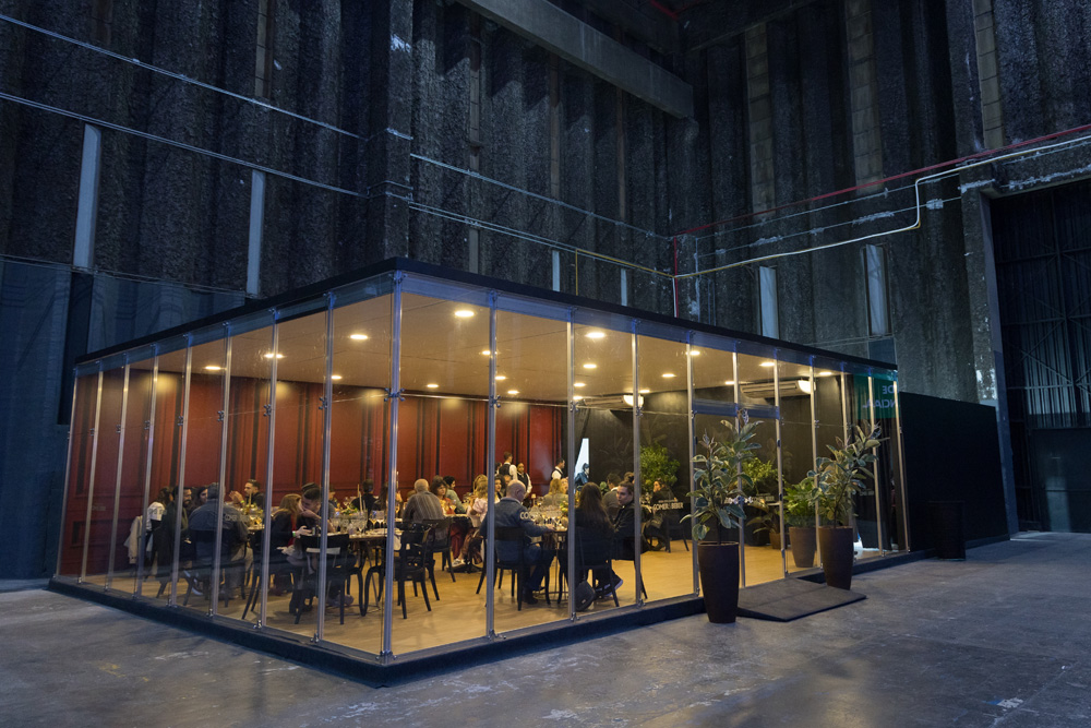 A imagem mostra uma sala fechada vista por fora com paredes de vidro. Do lado de dentro é possível ver mesas e cadeiras e pessoas sentadas.