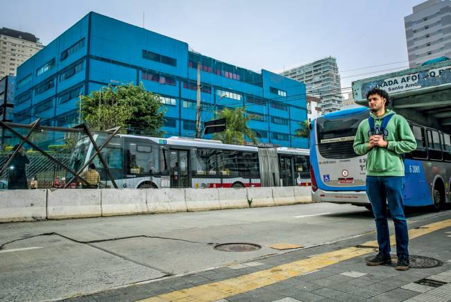 Imagem mostra jovem em ponto de ônibus olhando para a frente. Dois ônibus passam atrás do rapaz. Ele segura uma mochila