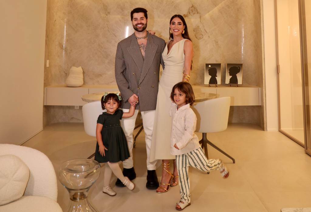 Mulher de cabelos longos posa de vestido branco ao lado de marido, com barba e sorriso, vestindo paletó cinza. Em frente aos dois estão duas crianças, um menino e uma menina. Todos posam em um grande ambiente de tons brancos e beges.