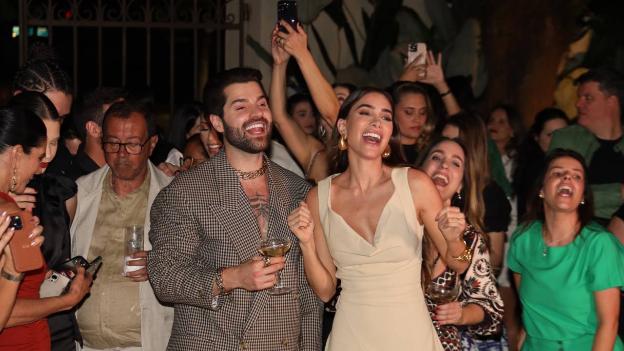 Homem de barba e moça de cabelos lisos longos sorriem e comemoram com champanhe na mão. Ele veste paletó cinza e ela um vestido bege. Pessoas aparecem ao fundo.