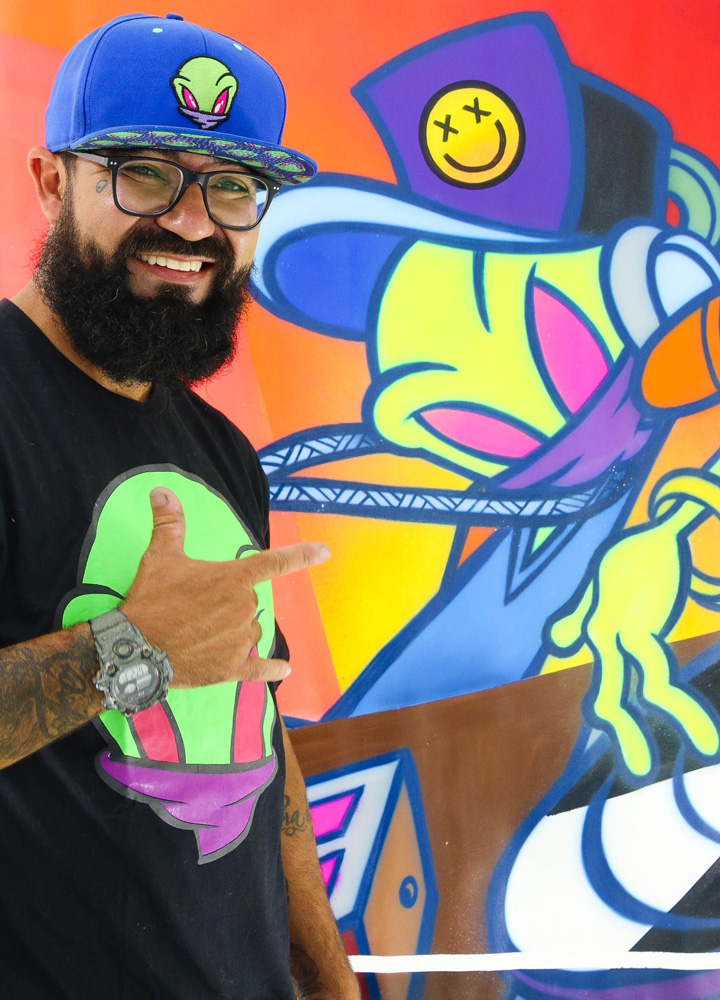 Homem branco de barba grande, óculos e boné posa com uma mão erguida em frente a parede de grafite colorido com um extraterrestre de boné.