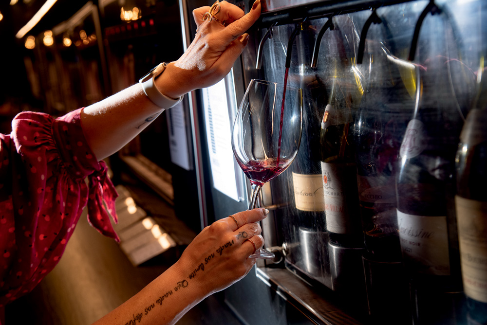 Mão servindo vinho tinto em máquina automática com garrafas dentro.
