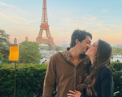 Imagem mostra rapaz beijando bochecha de moça em local que, ao fundo, aparece a Torre Eiffel, em Paris, França