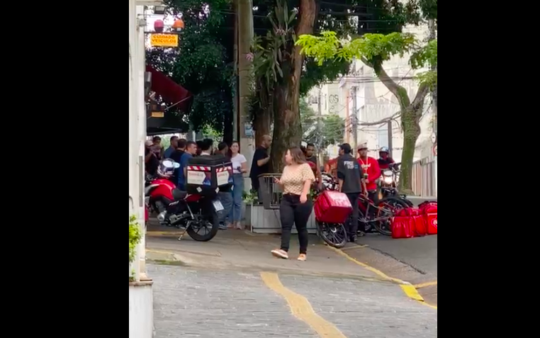 Frame de vídeo mostra pessoas passando na calçada de rua. Ao fundo, parcialmente encoberto por árvores, entregadores discutem com homem branco