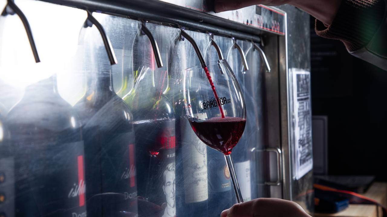 Imagem mostra garrafas de vinho dispostas dentro de máquina, com torneiras saindo da máquina, que é envidraçada. Uma das torneiras joga vinho em uma taça, segurada por uma pessoa