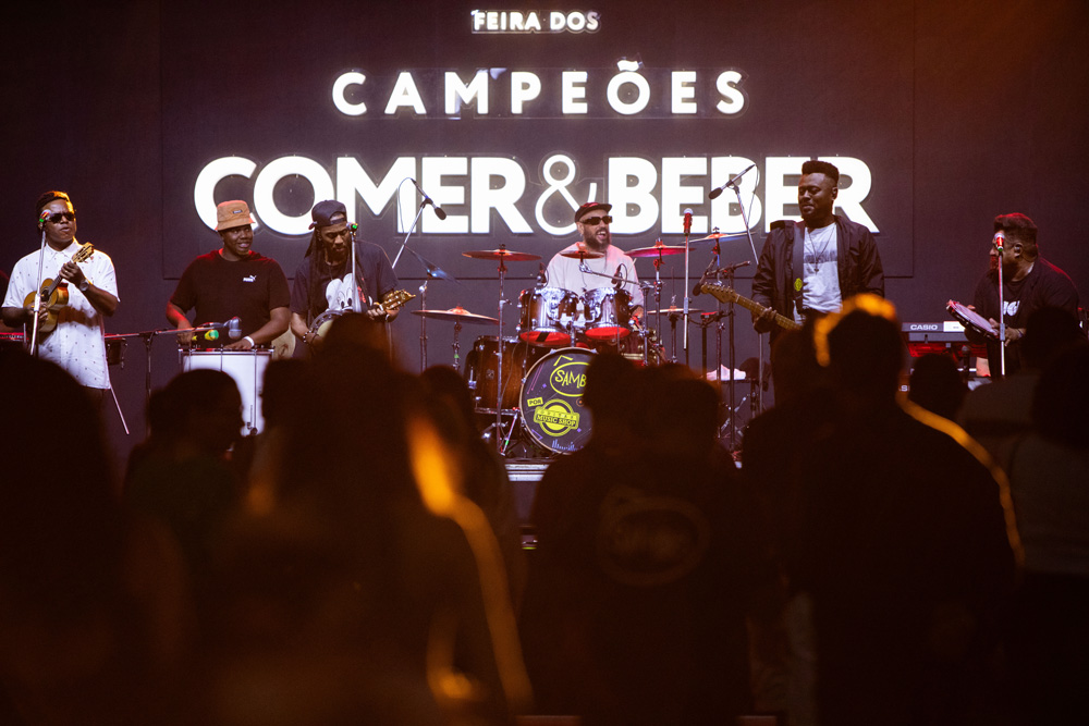 De frente, o palco com os músicos em cima. O do meio toca bateria, os dois à direita tocam guitarra e pandeiro. Os três a esquerda tocam banjo, tambor e cavaquinho.