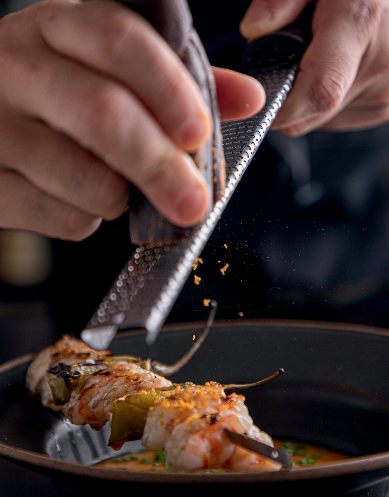 Espetinho de camarão apoiado sobre borda de prato fundo preto com mão ralando bottarga por cima.