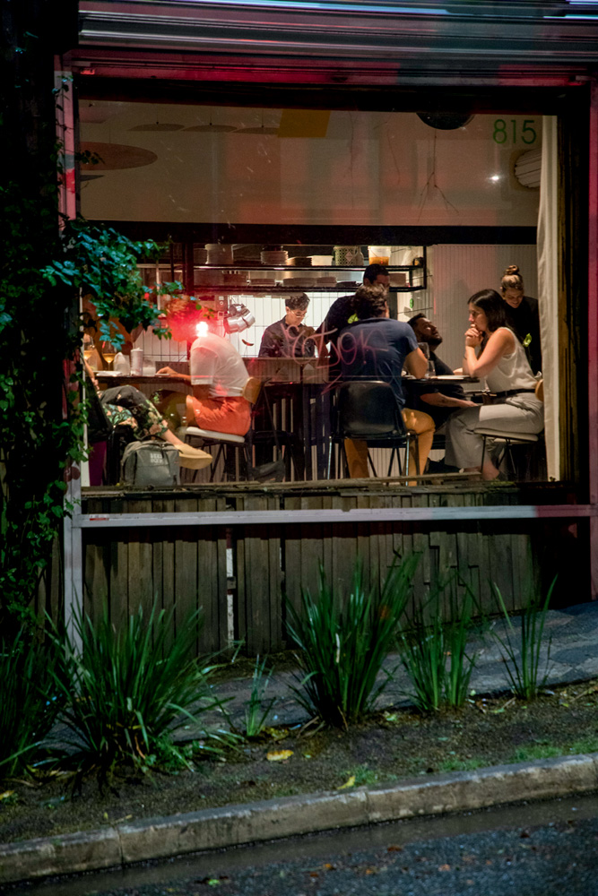 Fachada de restaurante com plantas na calçada, janela de vidro e pessoas sentadas em mesas do lado de dentro.