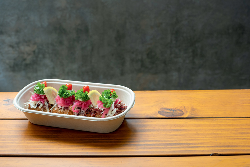 Em uma mesa de madeira, um pote bege com macaxeira e carne seca com cebola roxa, folhas verdes e tomate por cima.