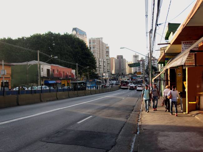 Imagem mostra avenida sem árvores e com fiação aparente. Duas faixas estão em foco e pessoas passam em calçada apertada