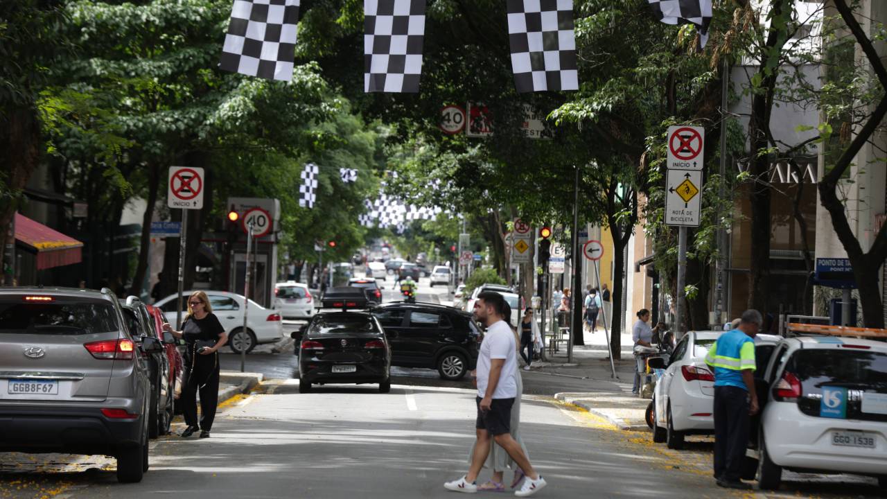 Bandeiras quadriculadas espalhadas pela rua Oscar Freire, nos Jardins, e uma pessoa atravessando a rua. Carros aparecem estacionados dos dois lados.