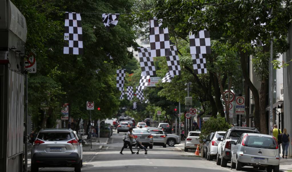 Bandeiras quadriculadas espalhadas pela rua Oscar Freire, nos Jardins, e uma pessoa atravessando a rua. Carros aparecem estacionados dos dois lados.