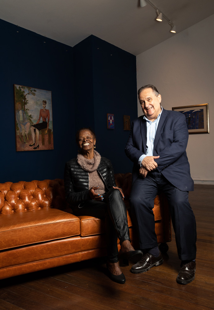 LEILAOVOLPI26 Sentados em sofá de couro marrom claro, uma mulher negra e um homem branco sorriem. Ela veste calça preta e jaqueta de couro preto, ele um terno azul com camisa azul claro. Ao fundo, um quadro grande retrata uma mulher. A obra está pendurada em uma parede de tom azul marinho.