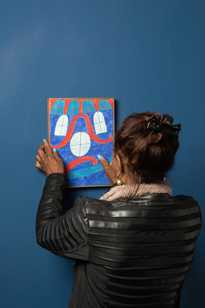 De costas, mulher negra vestida de jaqueta de couro pendura um quadro que retrata janelinhas e um fundo azul. A parede tem tom azul marinho.