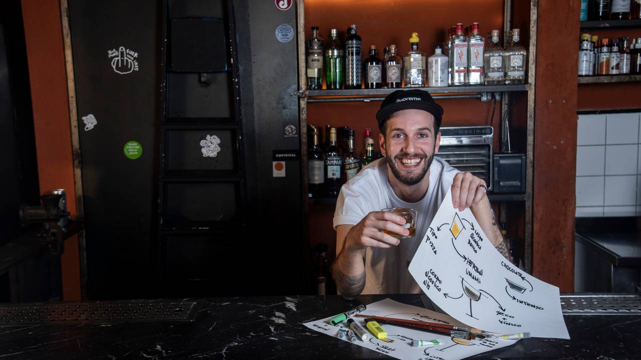 Homem de camiseta branca e boné sorrindo atrás de balcão de bar segurando um drinque na mão direita e desenhos de drinks na mão esquerda.
