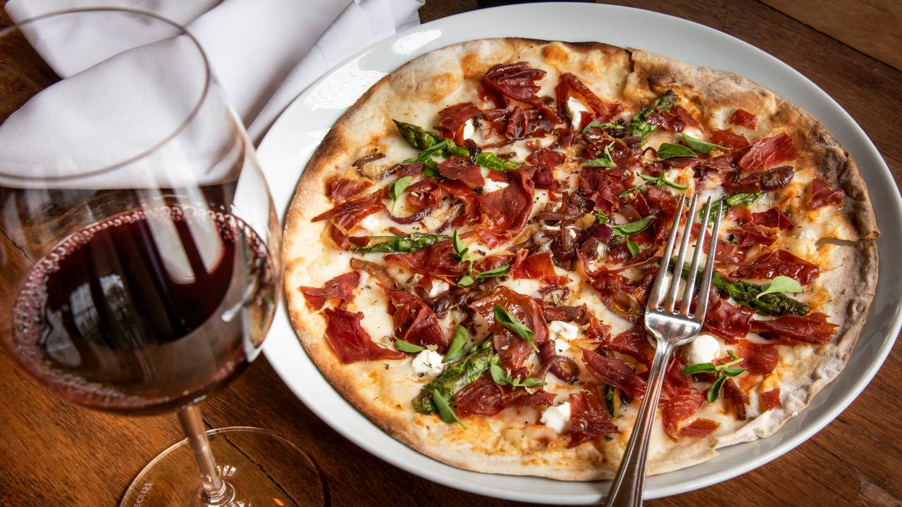 Imagem mostra uma pizza com fatias de presunto e pedaços de aspargos, ao lado, uma taça de vinho tinto.