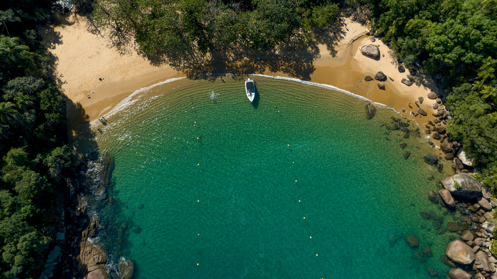 Imagem aérea mostra praia de águas verde clara com barco próximo da areia. Local tem grande volume de vegetação