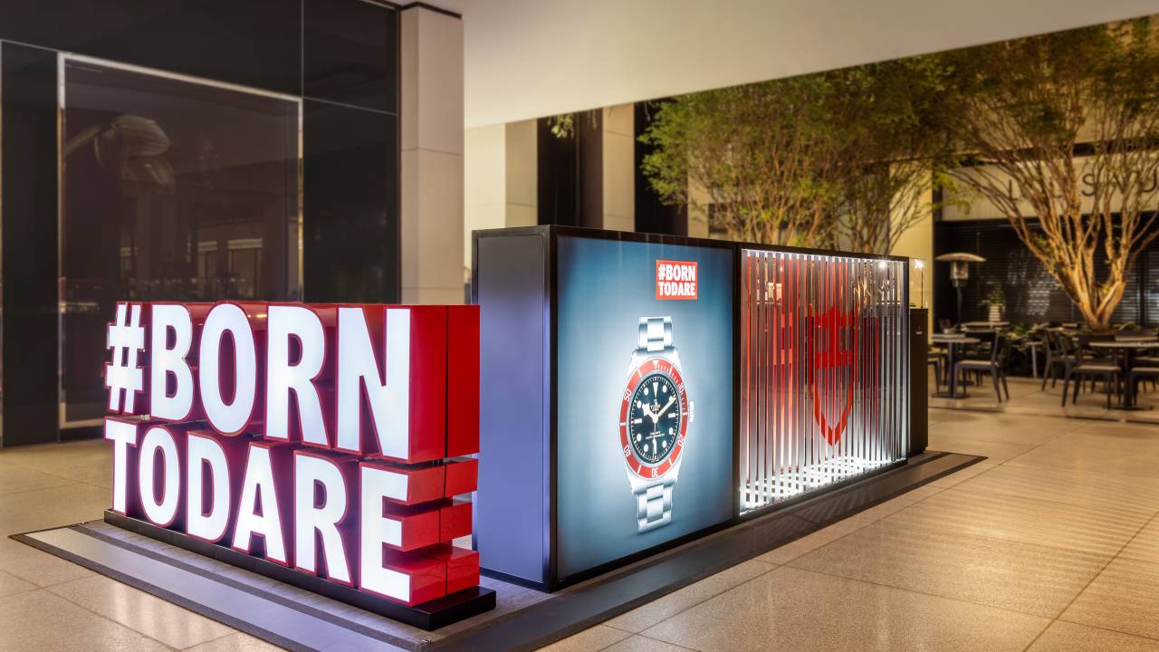 Com hashtag gigante "Born to Dare", foto exibe estande de loja de relógios no corredor de um shopping.