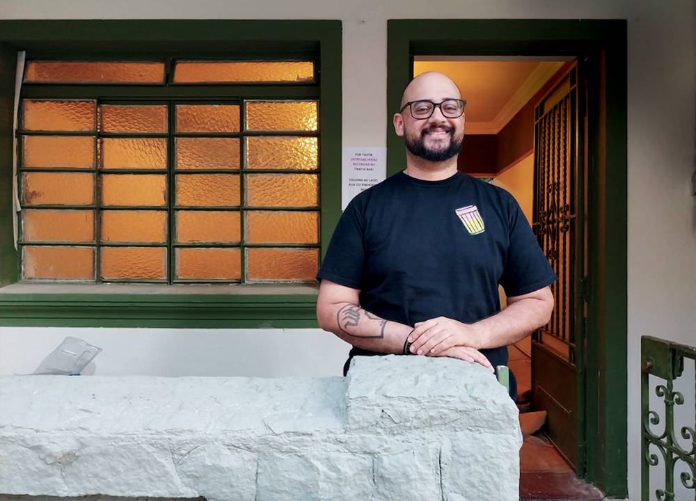 Retrato de Marco Aurélio Sena sorrindo, com camiseta preta e óculos na frente de uma casa com porta verde.