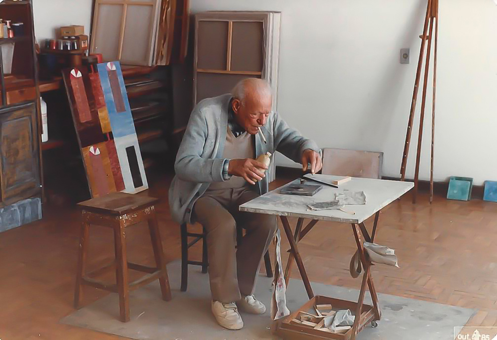 Foto exibe senhor grisalho e careca sentado e inclinado em mesa de desenho, com móveis espalhados ao fundo.