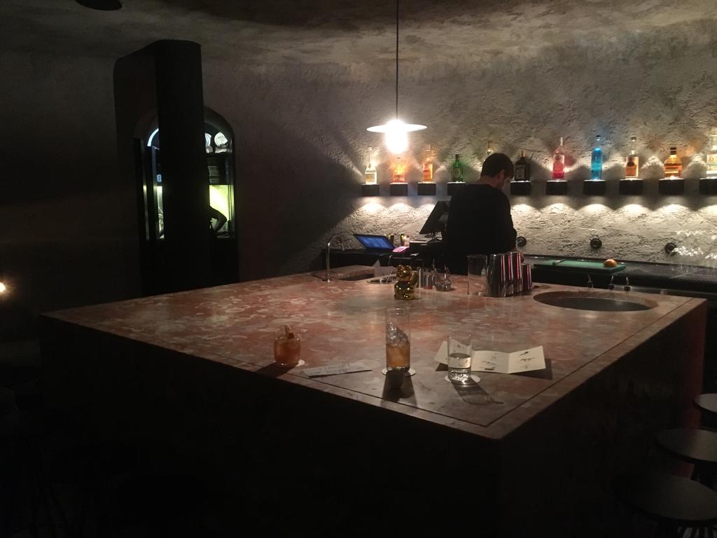 Toca da Raposa, em Lisboa Ambiente escuro com apenas uma lâmpada iluminando o centro. Embaixo dela uma grande mesa quadrada com alguns copos de drinques em cima.
