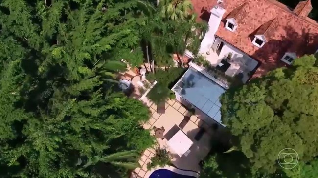 Imagem aérea mostra piscina rodeada de árvores com casa grande ao fundo
