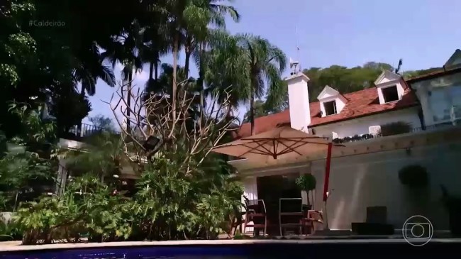Imagem mostra guarda-sol em primeiro plano e, ao fundo, grande casa, rodeada de árvores