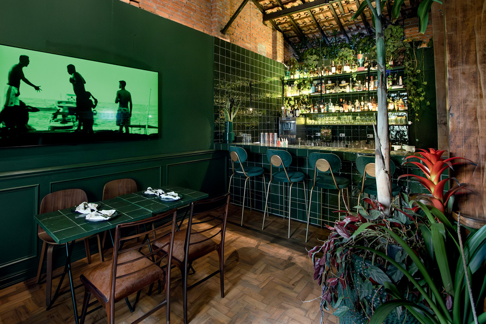 Ambiente com poucas mesas e cadeiras, parede verde escura e um balcão ao fundo.