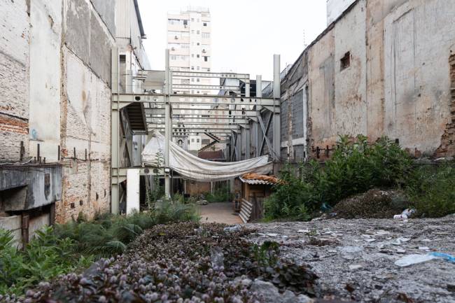 Imagem mostra terreno vazio, com mato nas bordas e ao fundo, uma estrutura metálica abandonada. Local é rodeado de prédios e, ao fundo, é possível ver parcialmente a torre da capela