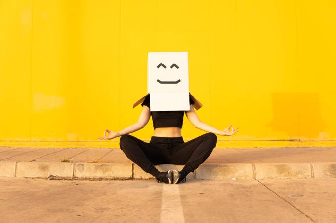 Woman wearing box with smiley face meditating in front of yellow wall on footpath