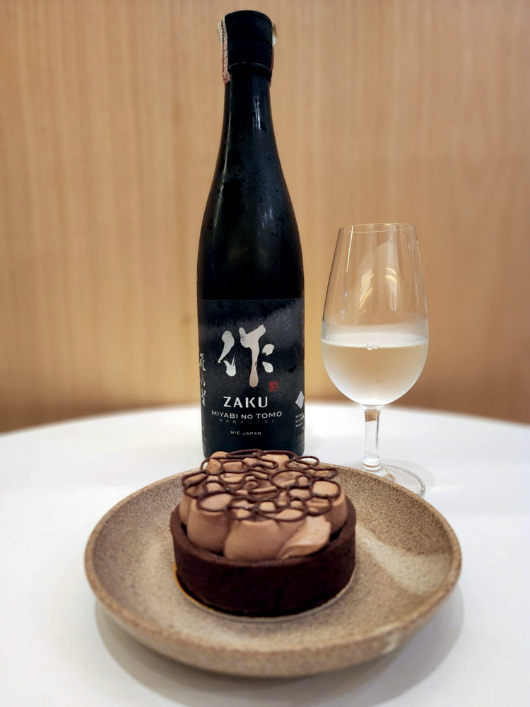 Circuito Comer & Beber – Amay tortinha de chocolate no prato fundo de cerâmica com garrafa preta de saquê atrás ao lado de dose em taça