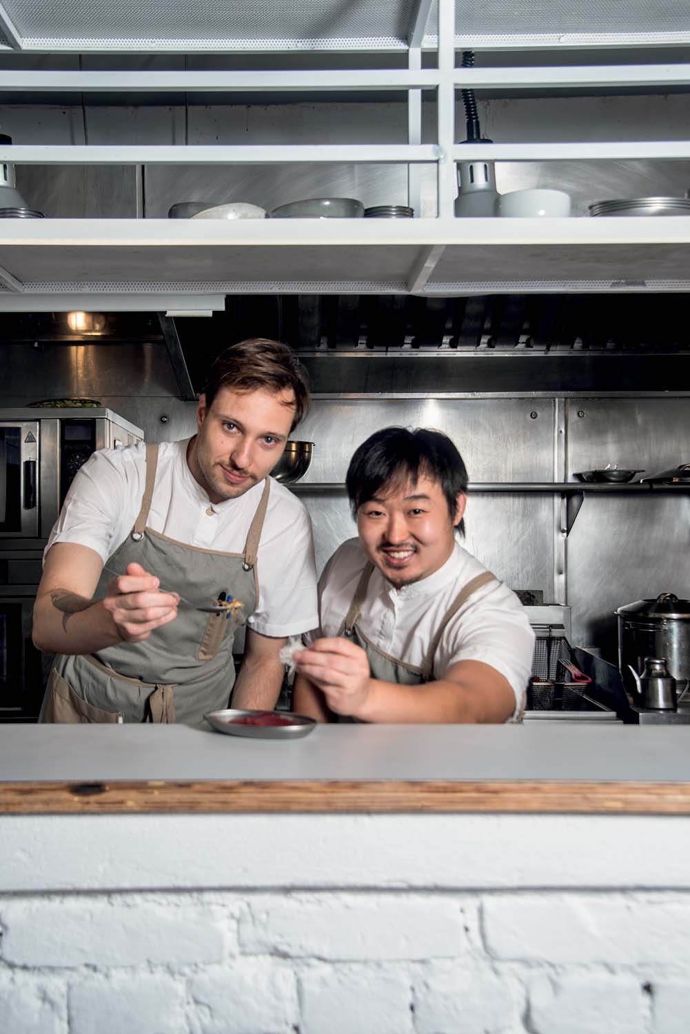 Dois chefs de camiseta branca coberta por avental cinza segurando colheres atrás de balcão em cozinha de restaurante