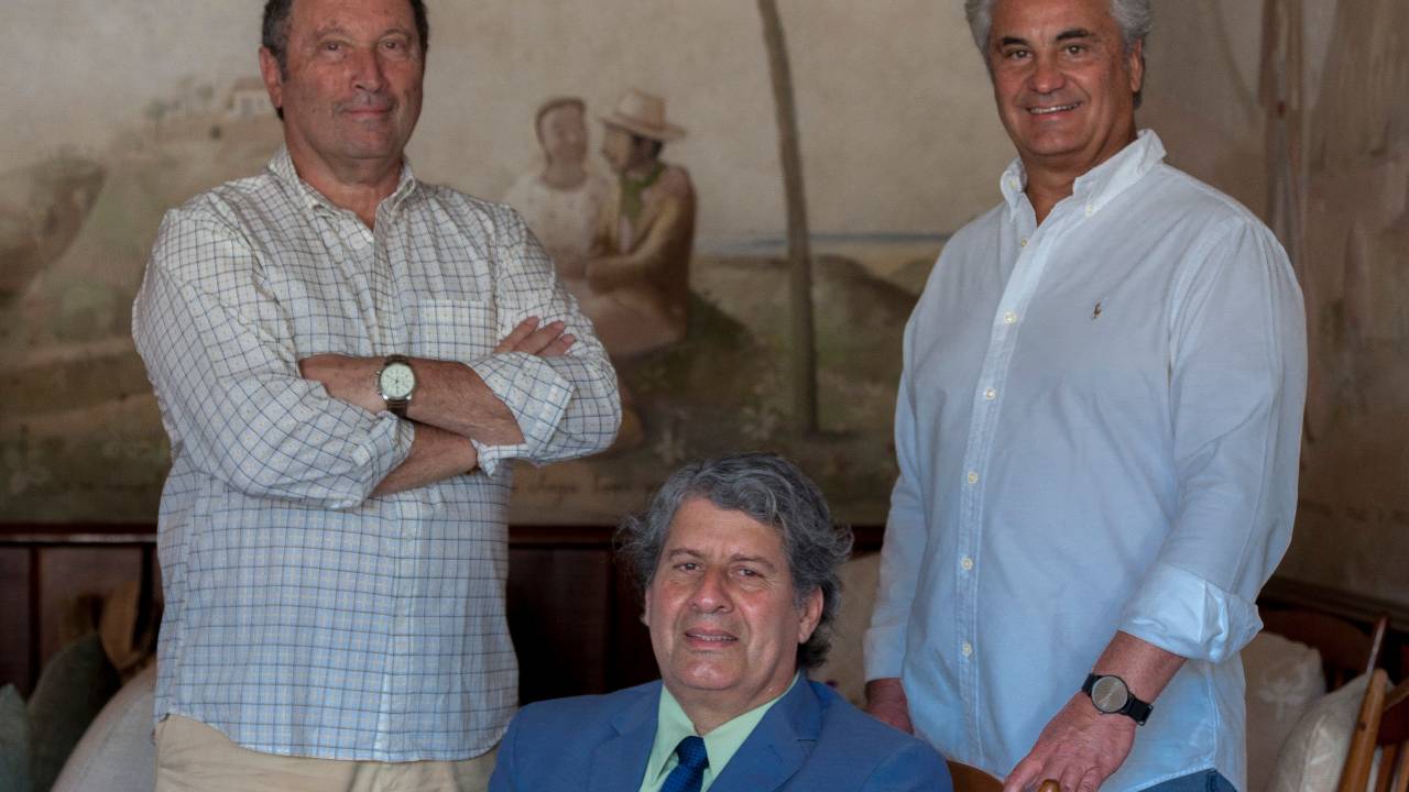 Três homens brancos posam sorrindo, dois de pé e um sentado, que veste terno azul. Os outros estão de camisa branca.