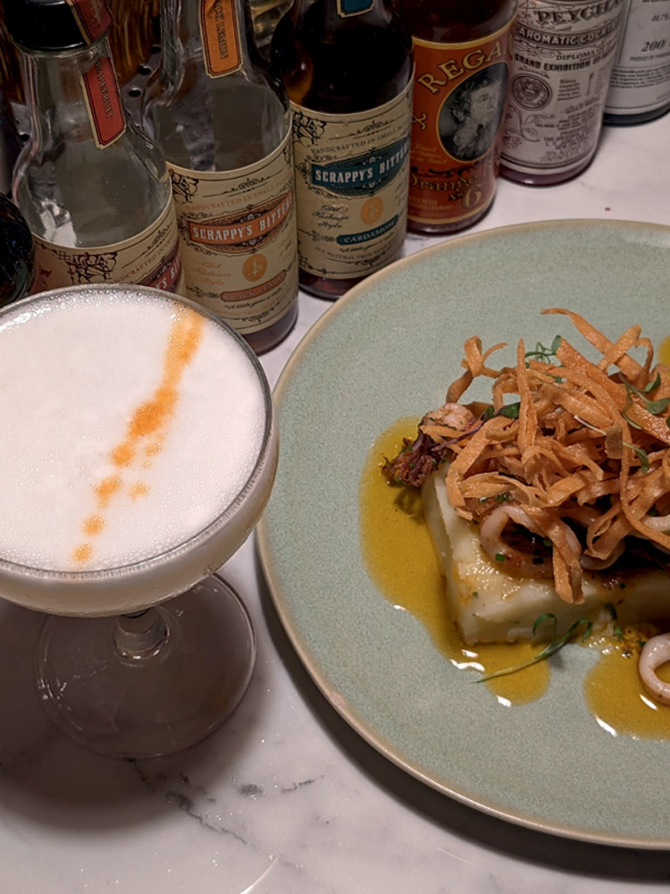 Circuito Comer & Beber – Cascasse Il Mondo Foto aérea de drinque com espuma branca com gotas laranjas ao lado de prato com peixe coberto de chips