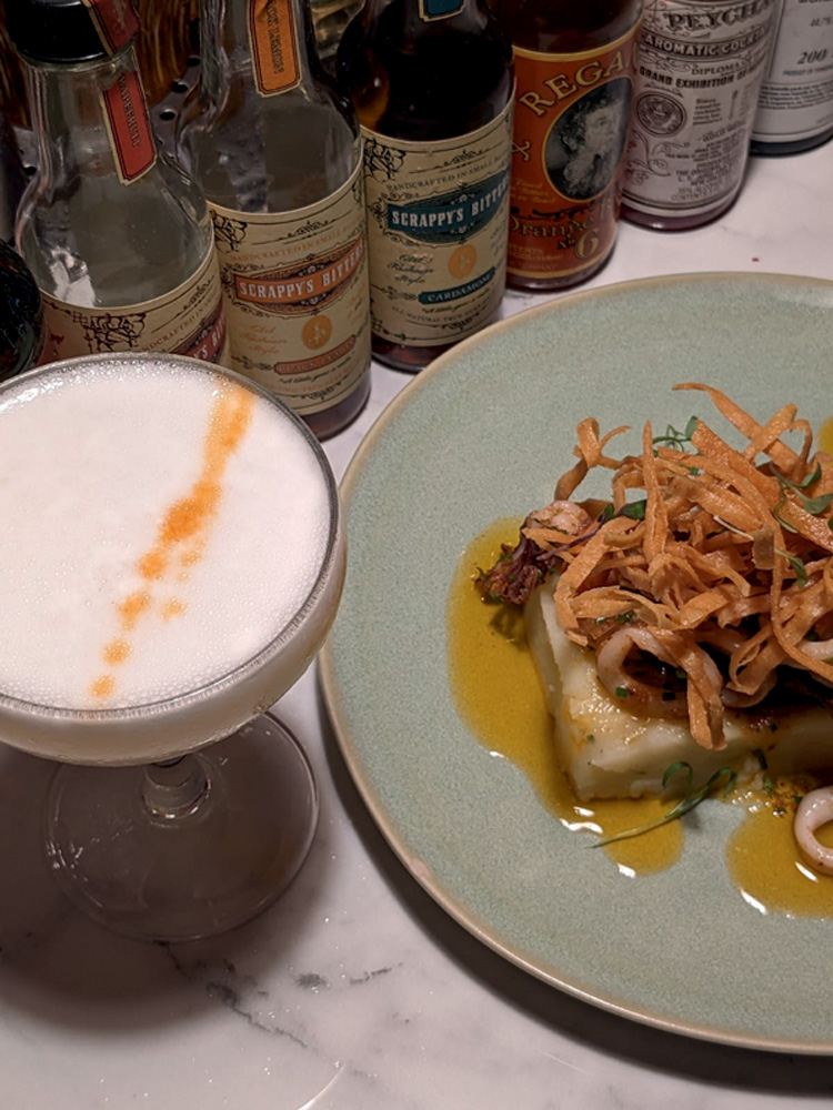 Foto aérea de drinque com espuma branca com gotas laranjas ao lado de prato com peixe coberto de chips