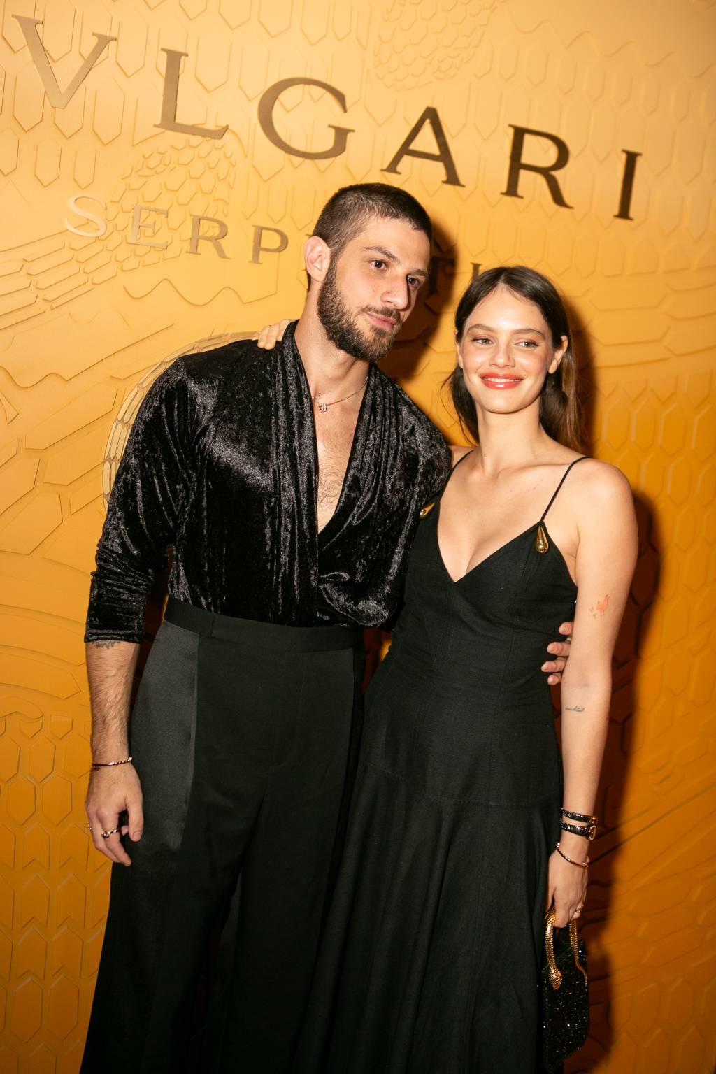 Casal posa em frente a parede laranja com a marca Bvlgari. Vestem peças pretas.