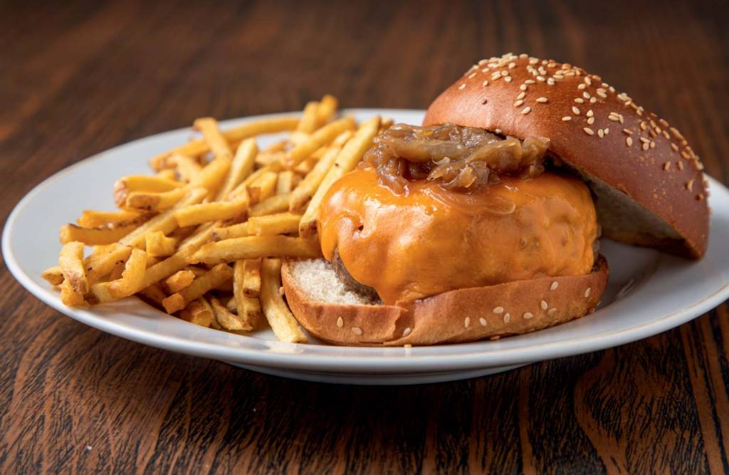 Sobre uma mesa de madeira há um prato branco com batatas fritas à esquerda e um hambúrguer com queijo cheddar e pão com gergelim do lado.
