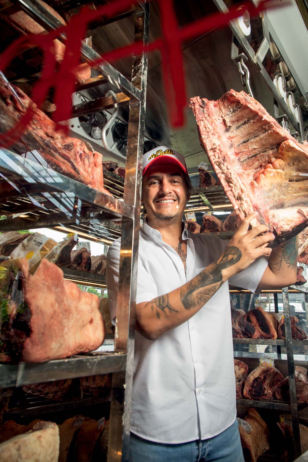 Um homem com avental branco e boné vermelho sorri para a foto e segura um pedaço grande de carne. A sua volta tem prateleiras com mais pedaços de carnes.