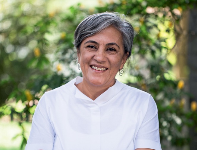 Mulher branca de cabelos grisalhos posa sorrindo com braços cruzados em frente a jardim. Veste camisa branca.