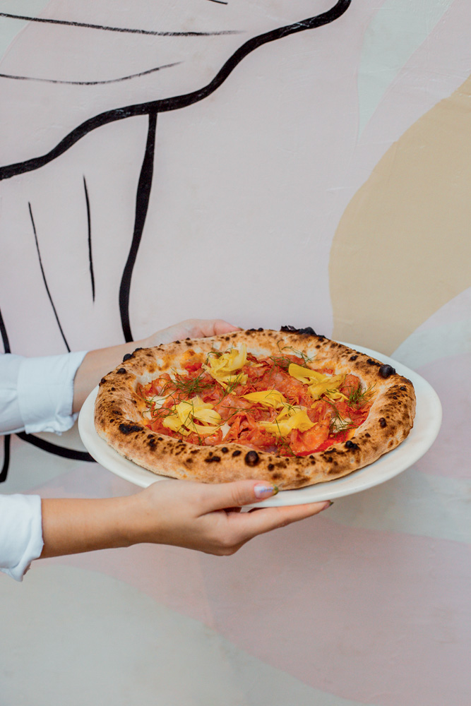 Mãos segurando prato de louça branca com pizza coberta por rodelas de linguiça