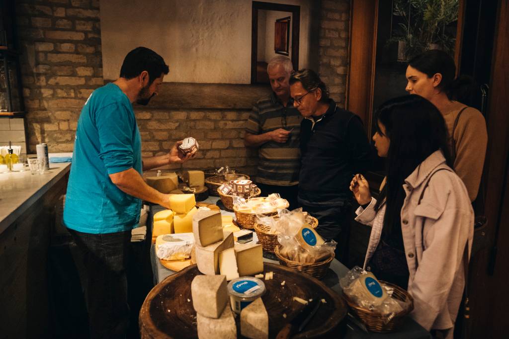 feira de queijos com mesa expositora ao centro, vendedor de camiseta azul à esquerda e público à direita.
