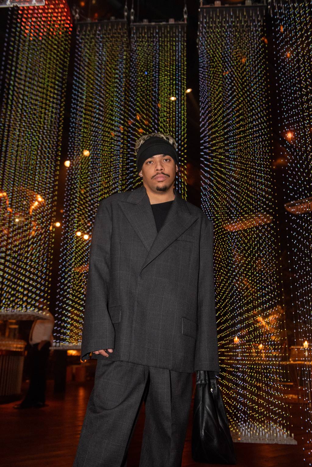Homem negro posa em frente a instalação de brilhantes suspensos e luminosos. Veste calça preta, blazer oversized preto e faixa preta na cabeça.
